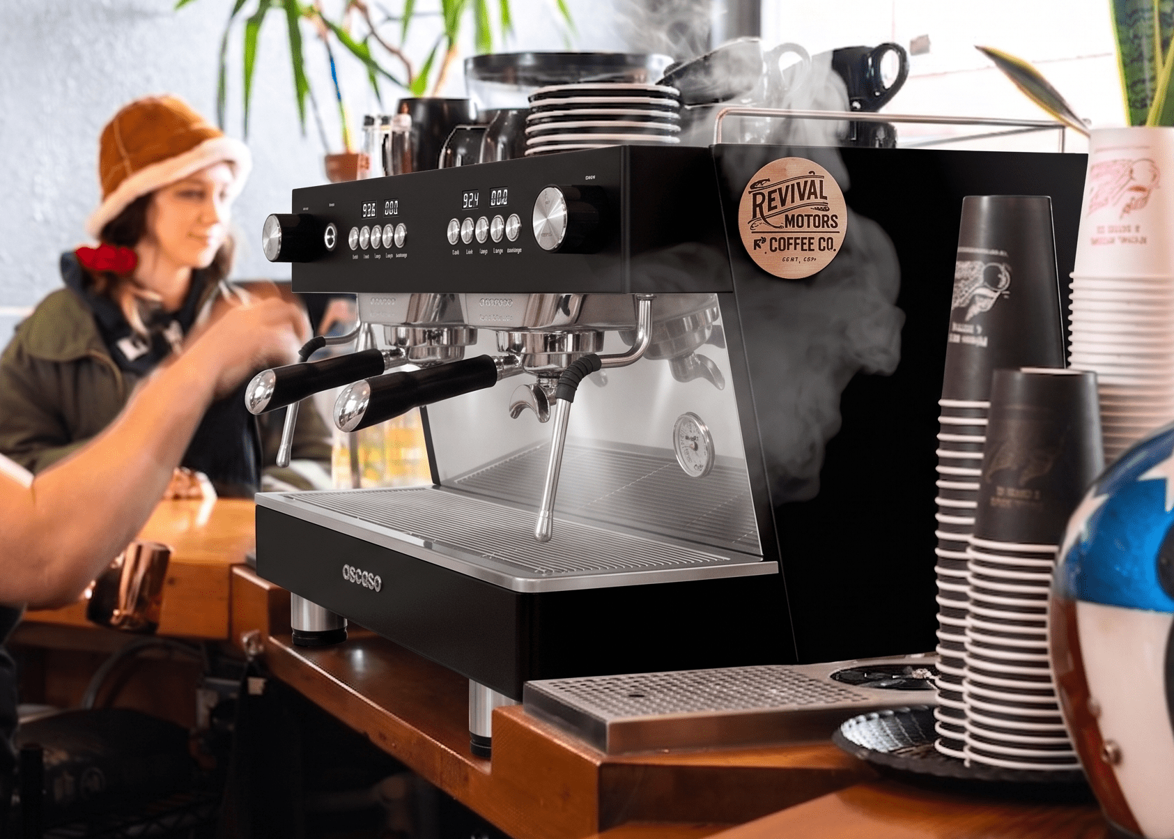 A black and stainless steel Revival Motors by Coffee Co. espresso machine on a wooden counter in a cafe setting, with a barista working in the background and stacks of cups nearby.